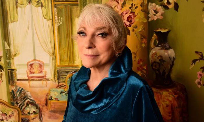 Judy Collins smiles in front of a flowery backdrop with vases and furniture.
